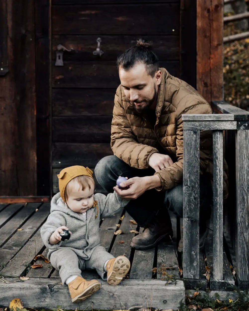 A dad in a work jacket and boots crouched on a porch with his baby